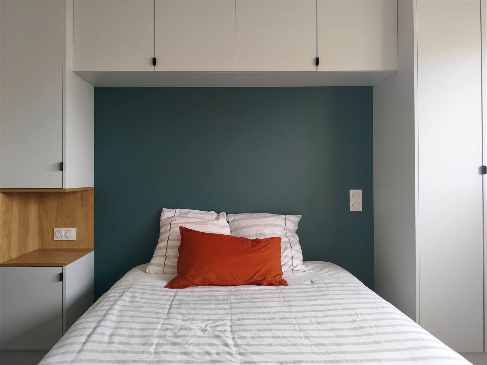 Le lit encadré de placards suspendus et d'un mur vert sapin, coussin terracotta