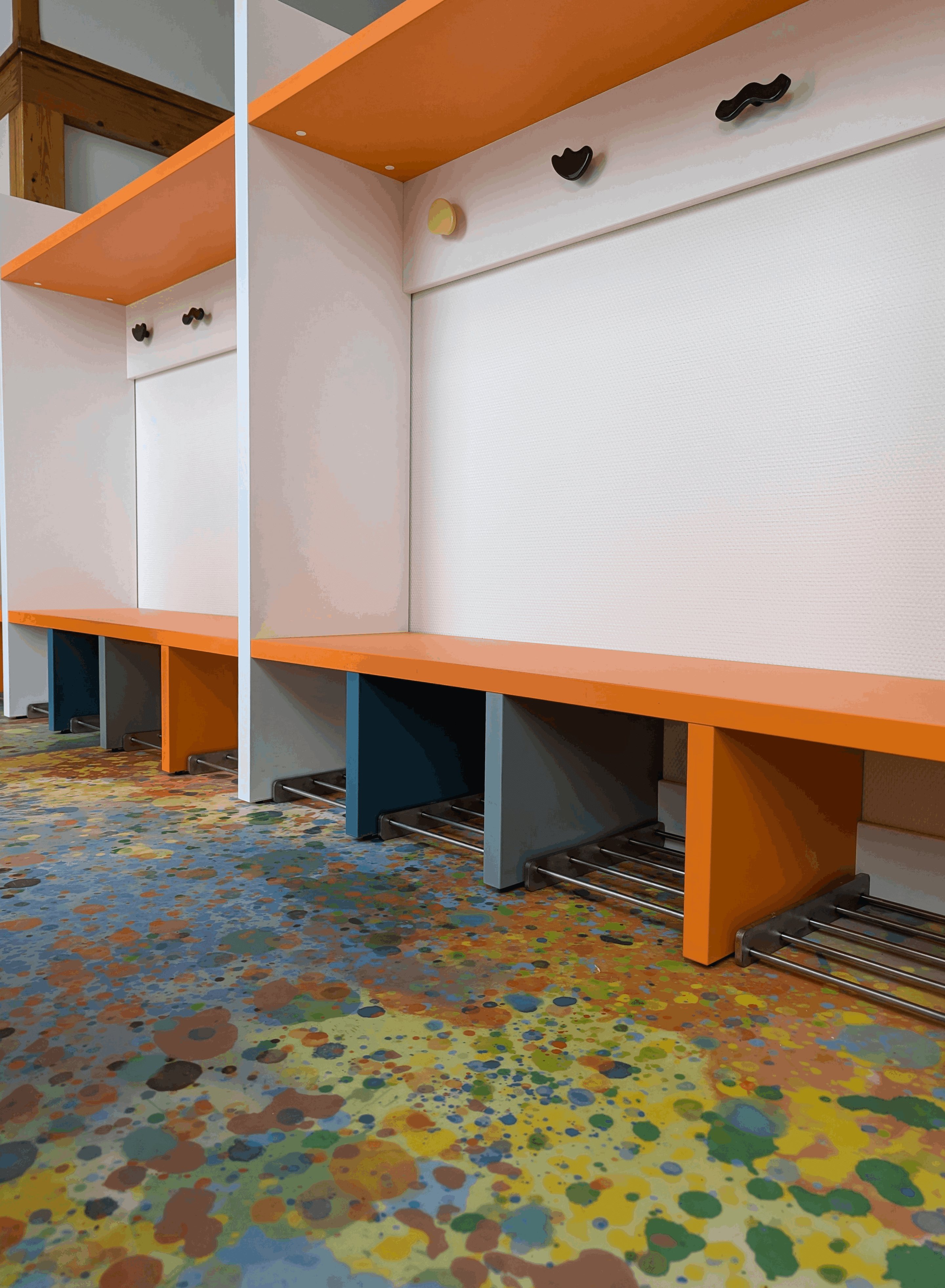 Banc orange et patères colorées sous l'étagère haute d'un vestiaire d'école maternelle
