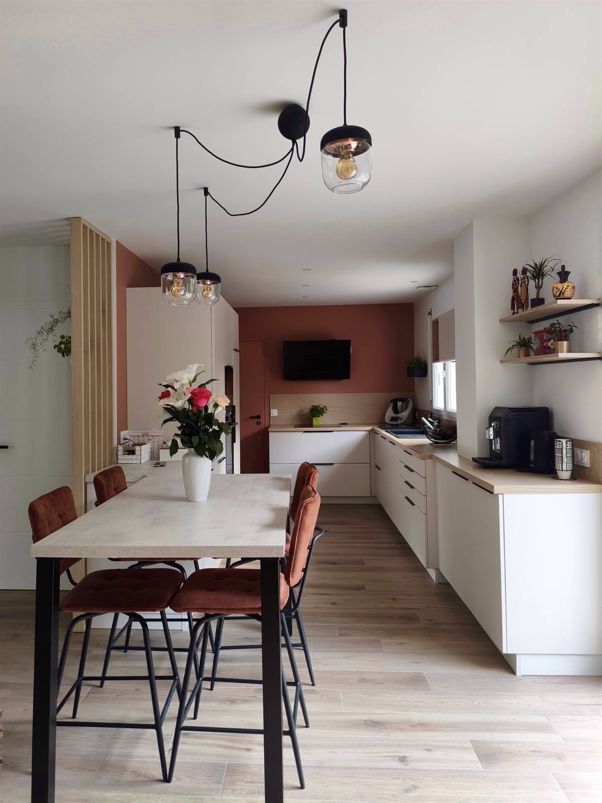 Vue depuis la table vers la cuisine : suspensions en verre, chaises cognac et mur terracotta
