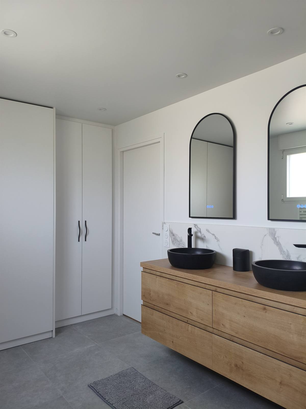 Salle de bain parentale à Groix : colonnes de rangement blanches, meuble double vasque en chêne et miroirs en arche