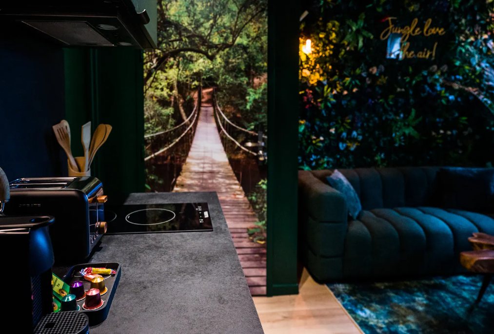 Cuisine ouverte sur le salon : plan de travail gris, papier peint panoramique de pont suspendu dans la jungle, canapé vert et mur végétal