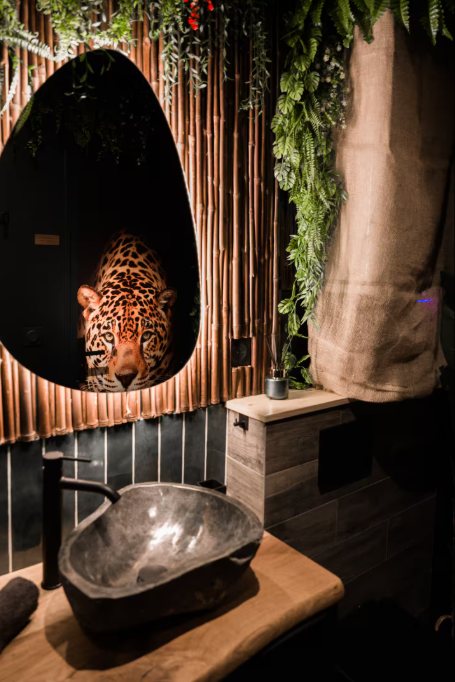 Salle de bains : vasque en pierre brute sur un plan en bois, miroir ovale au reflet de léopard et cloison en bambou