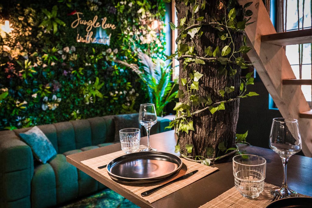 Table dressée contre un tronc d'arbre entouré de lianes, devant le canapé vert et le mur végétal au néon