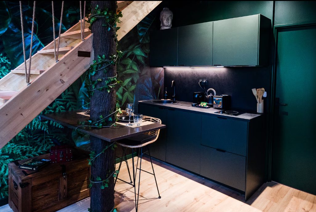 Cuisine vert foncé et noire sous l'escalier en bois, table haute fixée au tronc d'arbre et papier peint tropical