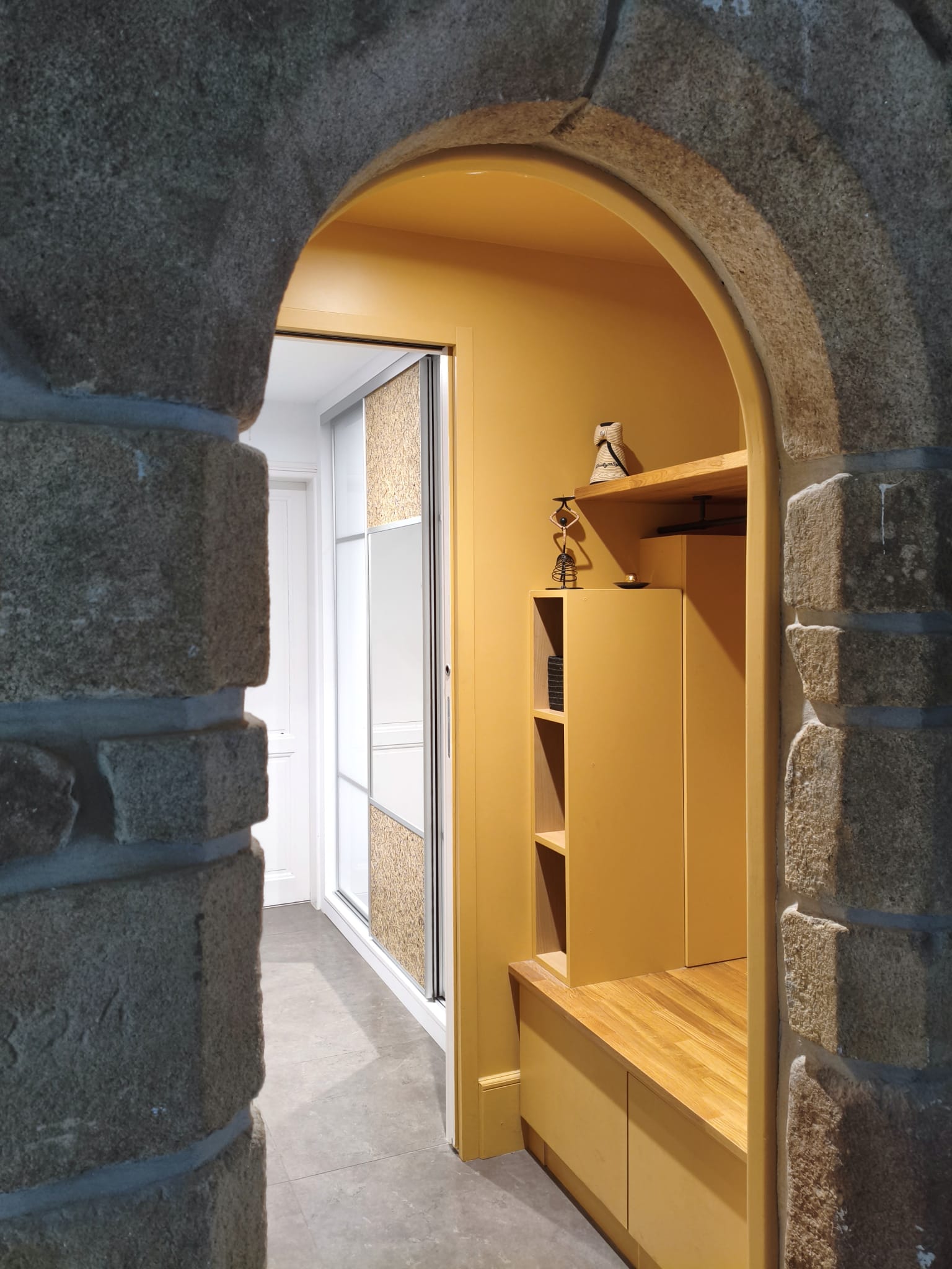 Sas d'entrée ocre vu à travers une arche en pierre, avec son meuble de rangement sur mesure