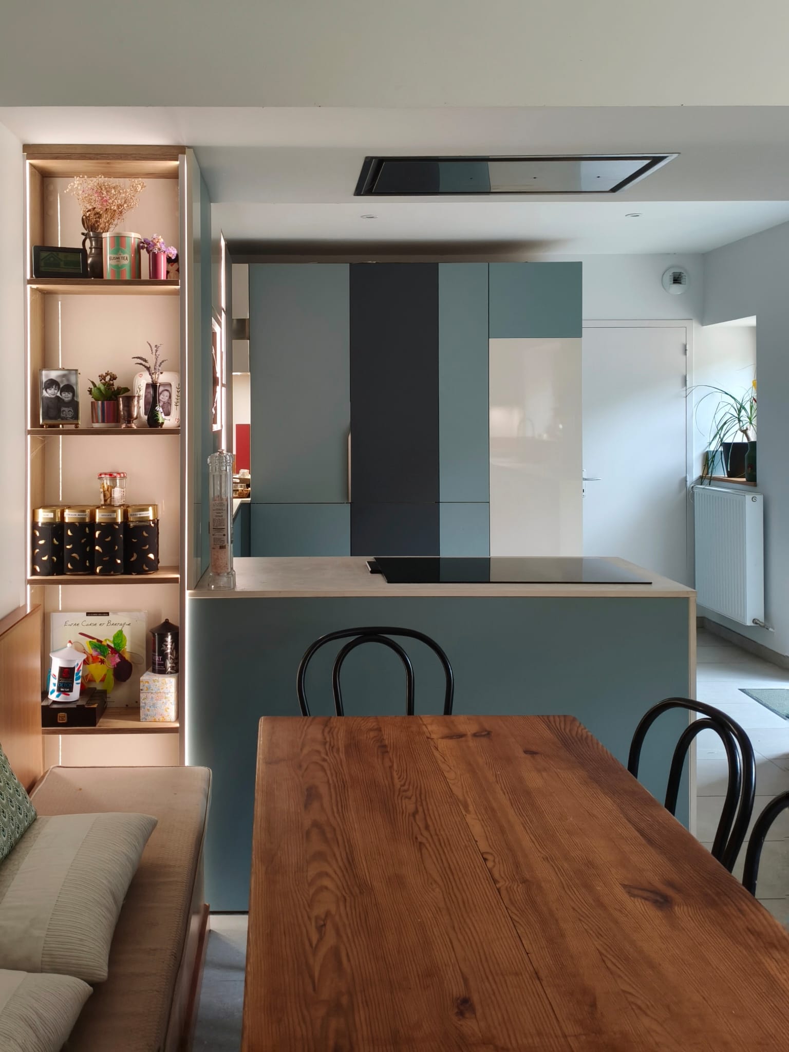 La cuisine vue depuis la table en bois : îlot, colonnes bicolores et étagères éclairées
