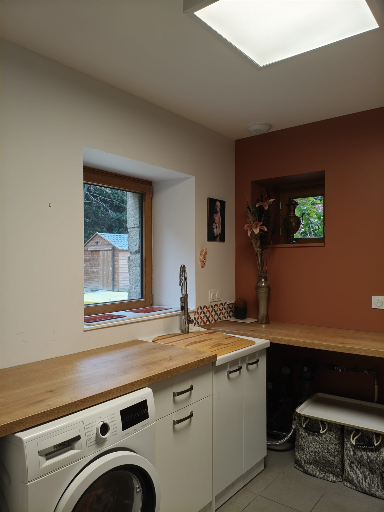 Buanderie : lave-linge sous un plan de travail en bois, meubles blancs et mur terracotta